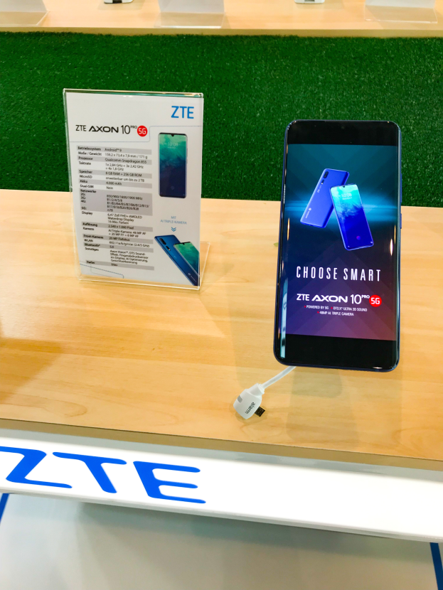 Ein ZTE Axon 10 Smartphone auf einem Tisch mit einem angeschlossenen Kabel, einer Tafel mit Text und anderen Gegenständen, mit dem Text "zte axon 10 in Indien gestartet" unten.