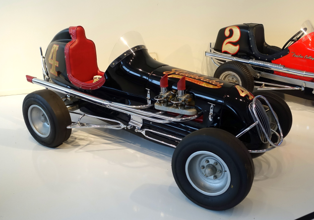 Zwei Oldtimer-Rennwagen in einem Museum ausgestellt, auf dem Boden stehend mit einer Wand im Hintergrund.