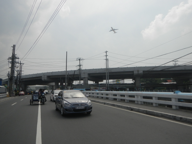 Ein Auto fährt auf einer Autobahn mit einer Brücke im Hintergrund, flankiert von Strommasten und Drähten, während ein Flugzeug darüberfliegt.