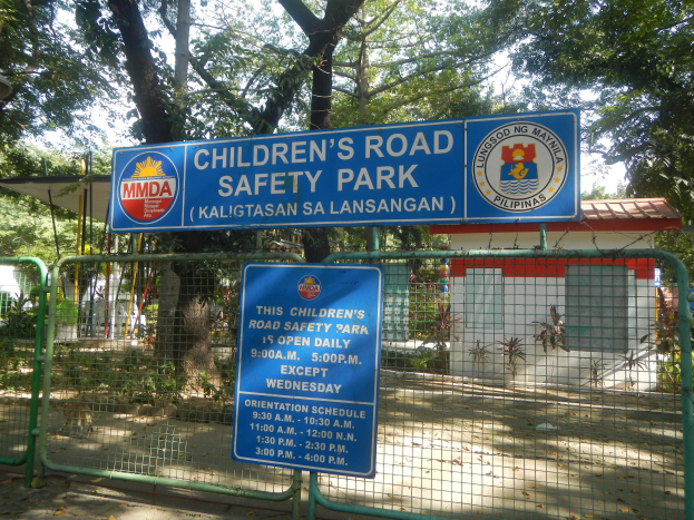 Kindersicherheitspark mit Metallzaun, zwei blauen Informationsschildern, einem kleinen Haus, Pflanzen, Bäumen und einer bewölkten Himmel