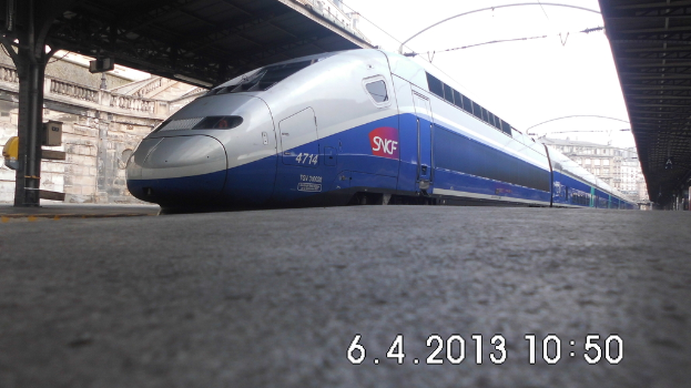 Ein Zug ist in der Mitte des Bildes zu sehen, mit einer Plattform und Text unten, Gebäuden und einem Dach links und Gebäuden, Dächern, Kabeln und Himmel oben.