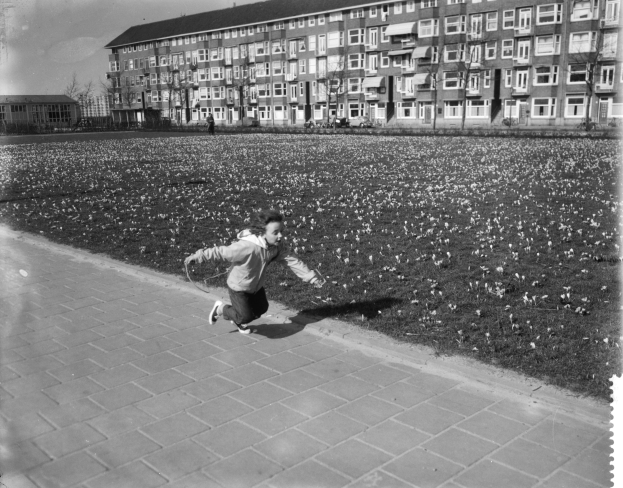 Ein junger Junge läuft über einen Gehweg in einem Park, umgeben von Gebäuden, Bäumen und einem klaren Himmel, trägt Schuhe.