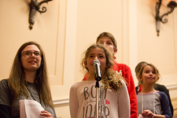 Eine Gruppe junger Mädchen steht nebeneinander vor einem Mikrofon, eine hält ein Blatt Papier in der Hand, mit an der Wand angebrachten Lampen im Hintergrund, die eine warme Atmosphäre schaffen.