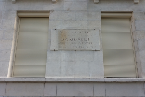 Ein zweistöckiges Gebäude mit zwei Fenstern mit Läden und einer Tafel an der Seite, wahrscheinlich ein Denkmal.