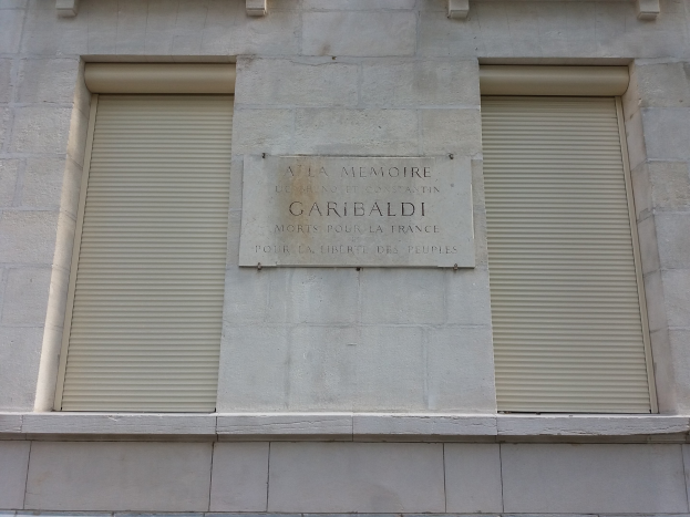 Ein zweistöckiges Gebäude mit zwei Fenstern mit Läden und einer Tafel an der Seite, wahrscheinlich ein Denkmal.