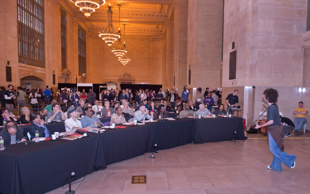 Ein Konzertszene mit einem Mann, der auf der rechten Seite Gitarre spielt und singt, einer Menge Menschen, einer Wand und Fenstern im Hintergrund, Lichtern oben und einem Boden unten.