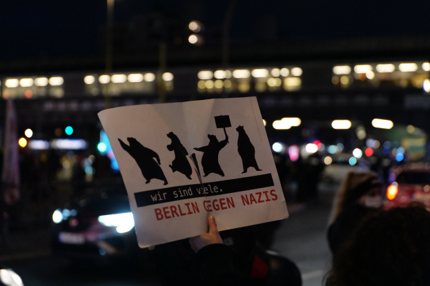 Person mit einer Tafel mit der Aufschrift "Berlin gegen Nazis" vor einer Menge und Fahrzeugen auf einer Straße, mit Gebäudebeleuchtung im Hintergrund.