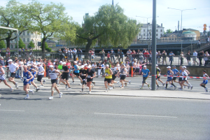Gruppe von Menschen, die bei einem Marathon auf einer Straße mit Absperrungen, Zuschauern und städtischer Umgebung unter einem bewölkten Himmel laufen.