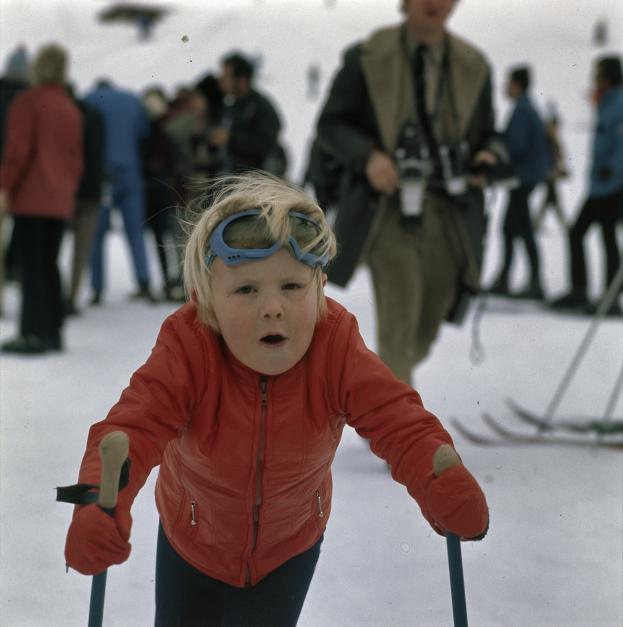 Ein junger Junge in roter Jacke und Brille, der eine schneebedeckte Hülle mit Ski-Stöcken hinabfährt, mit mehreren Personen und einigen, die Snowboards in der unscharfen Hülle halten.