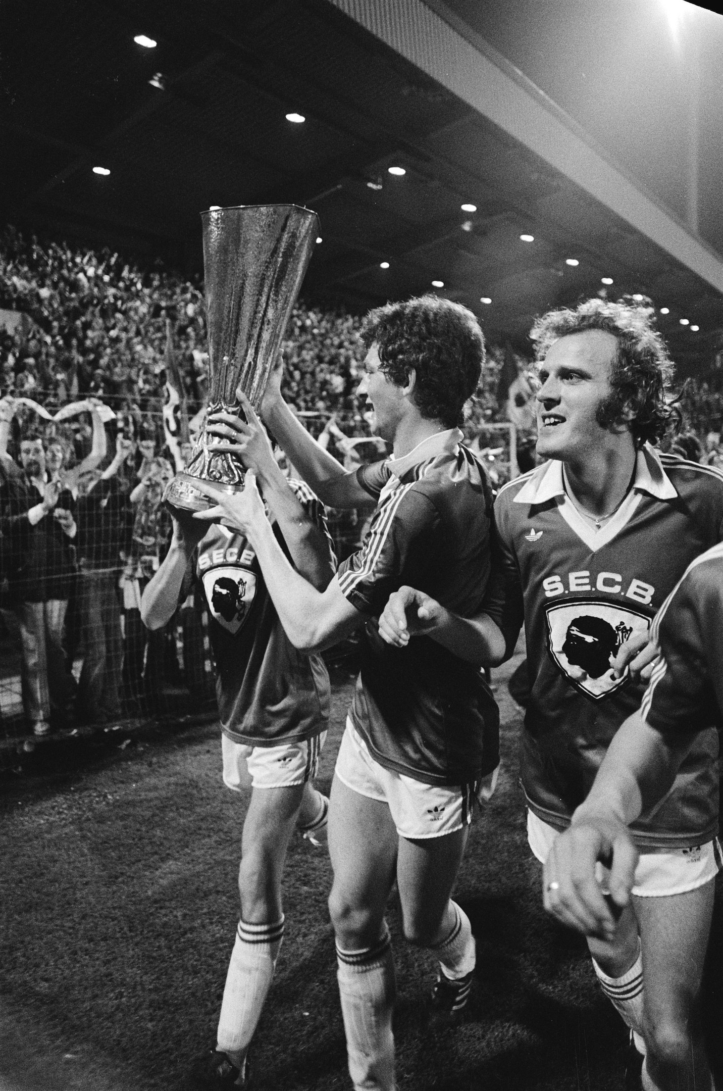 Schwarzes und weißes Bild von Fußballspielern, die mit einem Pokal auf einem Feld feiern, umgeben von einer Menge hinter einem Zaun unter Stadionbeleuchtung.