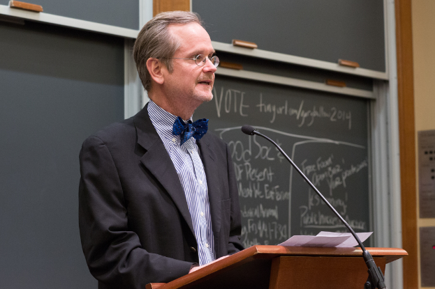 Ein Mann in einem schwarzen Anzug steht an einem Rednerpult mit Papieren, spricht in ein Mikrofon, mit einer Tafel im Hintergrund, auf der Text steht.