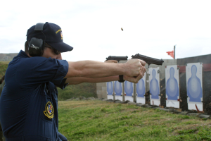 Ein Mann in einer blauen Uniform, Mütze, Brille und Kopfhörern zielt mit einer Pistole auf ein Ziel, das von Gras umgeben ist, mit einer Fahne und anderen Menschen im Hintergrund unter einem sichtbaren Himmel.
