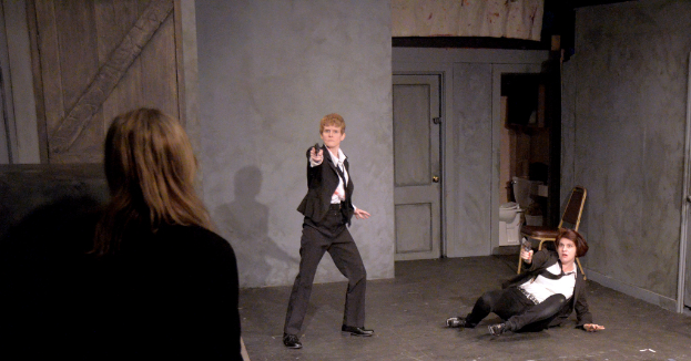 Zwei Frauen in schwarzen Anzügen, eine sitzend auf dem Boden eine Waffe haltend, die andere stehend und eine Waffe haltend, mit einer dritten Frau in der Nähe, in einem Raum mit einem Stuhl, einer Toilette und einer hölzernen Tür.