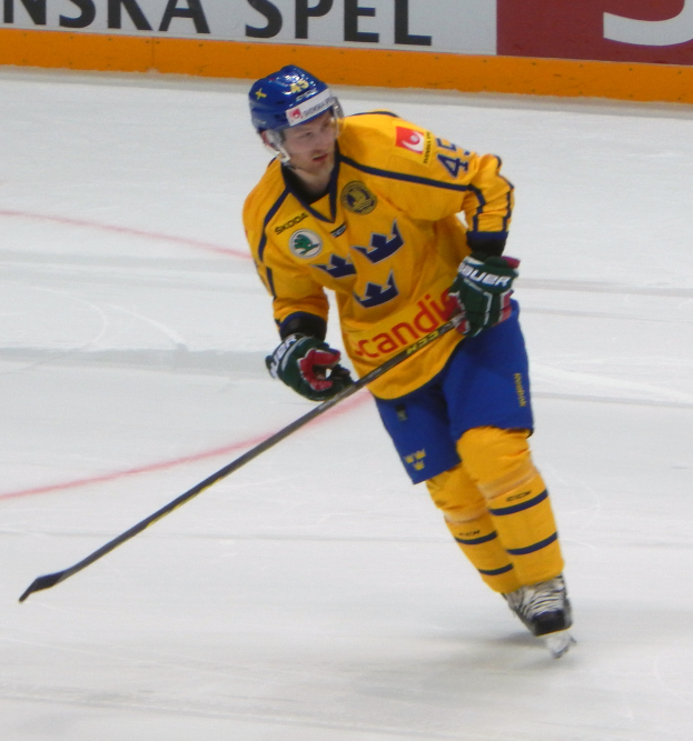 Ein Eishockeyspieler in Aktion auf dem Eis, mit Helm, Handschuhen und Schlittschuhen, der einen Hockeystock hölt, mit einer Tafel im Hintergrund.