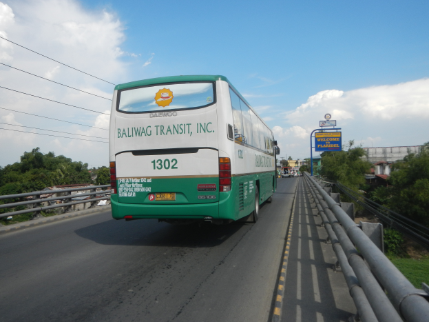 Ein grüner und weißer Bus fährt auf einer Autobahn mit Leitplanken auf beiden Seiten, mit Bäumen, Gebäuden, Texttafeln, Polen, Drähten und einem klaren blauen Himmel im Hintergrund.