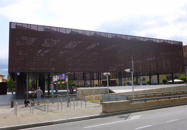Großes modernes Gebäude mit zahlreichen Fenstern als neuer Eingang zur Stadtmitte, umgeben von städtischer Infrastruktur wie Straßenlaternen, Fahrzeugen, Fußgängern und Bäumen unter einem bewölkten Himmel.