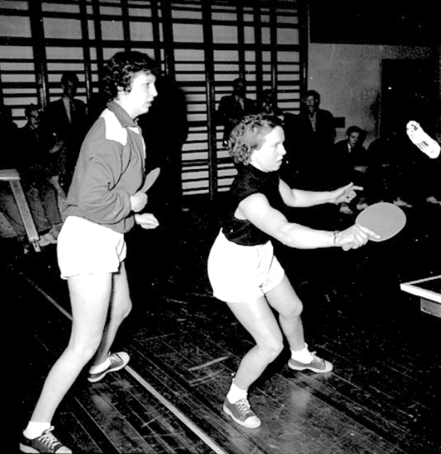 Zwei Frauen beim Tischtennis auf einem Court, jede hält einen Schläger in der Hand, mit einigen Zuschauern und einem Tischtennis-Tisch im Hintergrund, dargestellt in Schwarz-Weiß.
