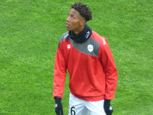 Ein junger Mann in einer roten Jacke und weißen Shorts steht auf einem Fussballfeld und schaut nach rechts mit einem entschlossenen Ausdruck, tragend Baseball-Handschuhe.