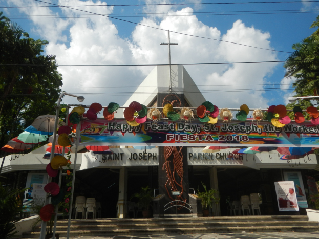Gebäude mit einem "Frohen Festtag St. Josef der Arbeiter"-Schild, Treppe zum Eingang, ein Bogen mit hängenden Dekorationen, Außenbestuhlung mit Stühlen und Tischen, Topfpflanzen, flankierende Bäume und ein bewölkter Himmel.