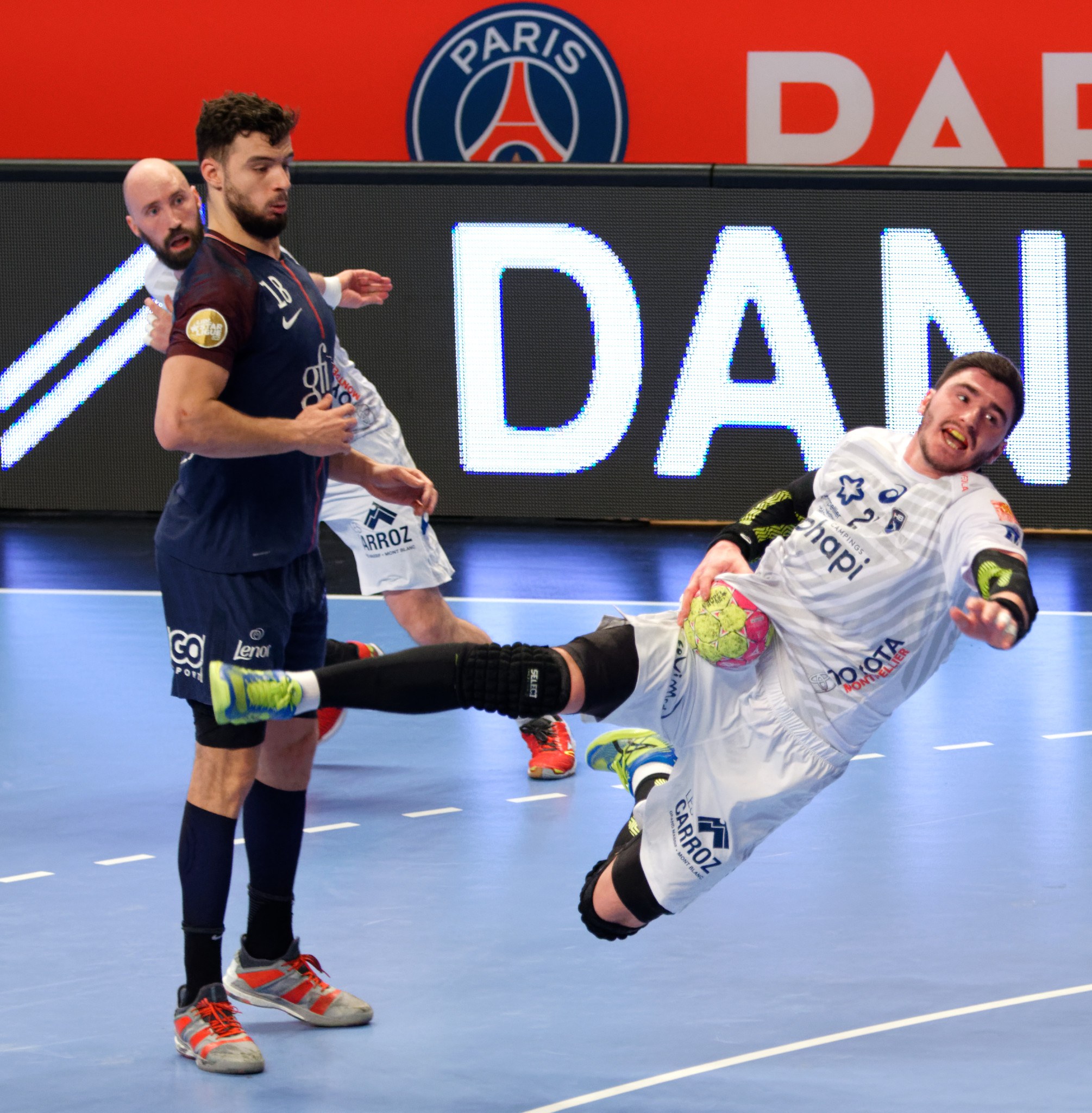 Eine Gruppe von Männern, die auf einem Handballfeld spielen, wobei einer den Ball hält, und ein Paris Saint-Germain-Futsal-Siegertafel im Hintergrund.