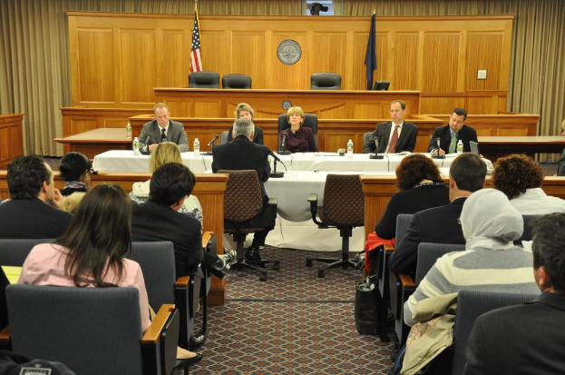 Ein Versammlungsraum mit einer Gruppe von Menschen, zwei fremden Flaggen, einem orangen Schreibtisch mit Wasserflaschen und einem Mikrofon.