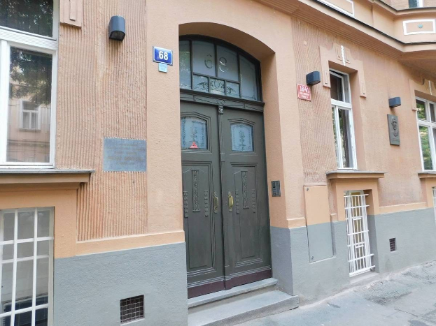 Ein zweistöckiges Gebäude mit einer grünen Tür und Fenstern an einer städtischen Straße, das einen Fußweg zum Gebäude und eine Schilderung für den Verkauf einer Wohnung zeigt.