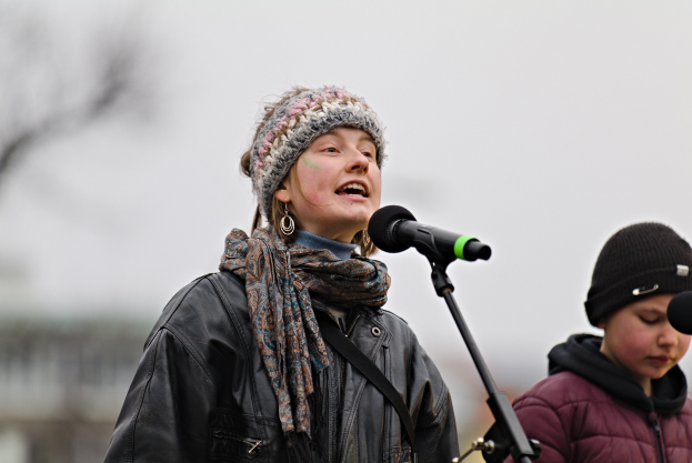 Eine Frau in einer schwarzen Jacke, einem Schal und einer Mütze singt in ein Mikrofon auf der Bühne, neben einer anderen Person in einer maronenfarbenen Jacke und einer schwarzen Mütze, während eine unscharfe Menge zusieht.