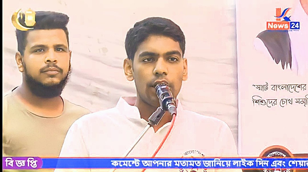 Zwei Männer stehen nebeneinander vor einem Fernseher, einer spricht in ein Mikrofon, der andere trägt ein graues T-Shirt und unterhält sich in einem scheinbar medialen Setting.