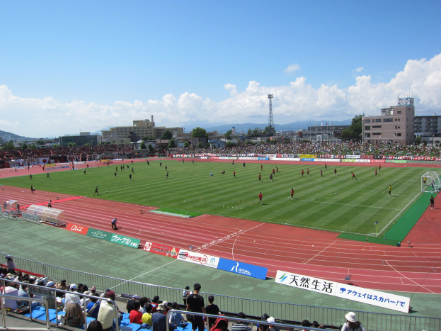 Großes Stadion voller Zuschauer bei einem Fußballspiel, mit Sitzplätzen und Stehplätzen, Geländern, Beschilderung und umliegenden Gebäuden, Bäumen, Hügeln und bewölktem Himmel.