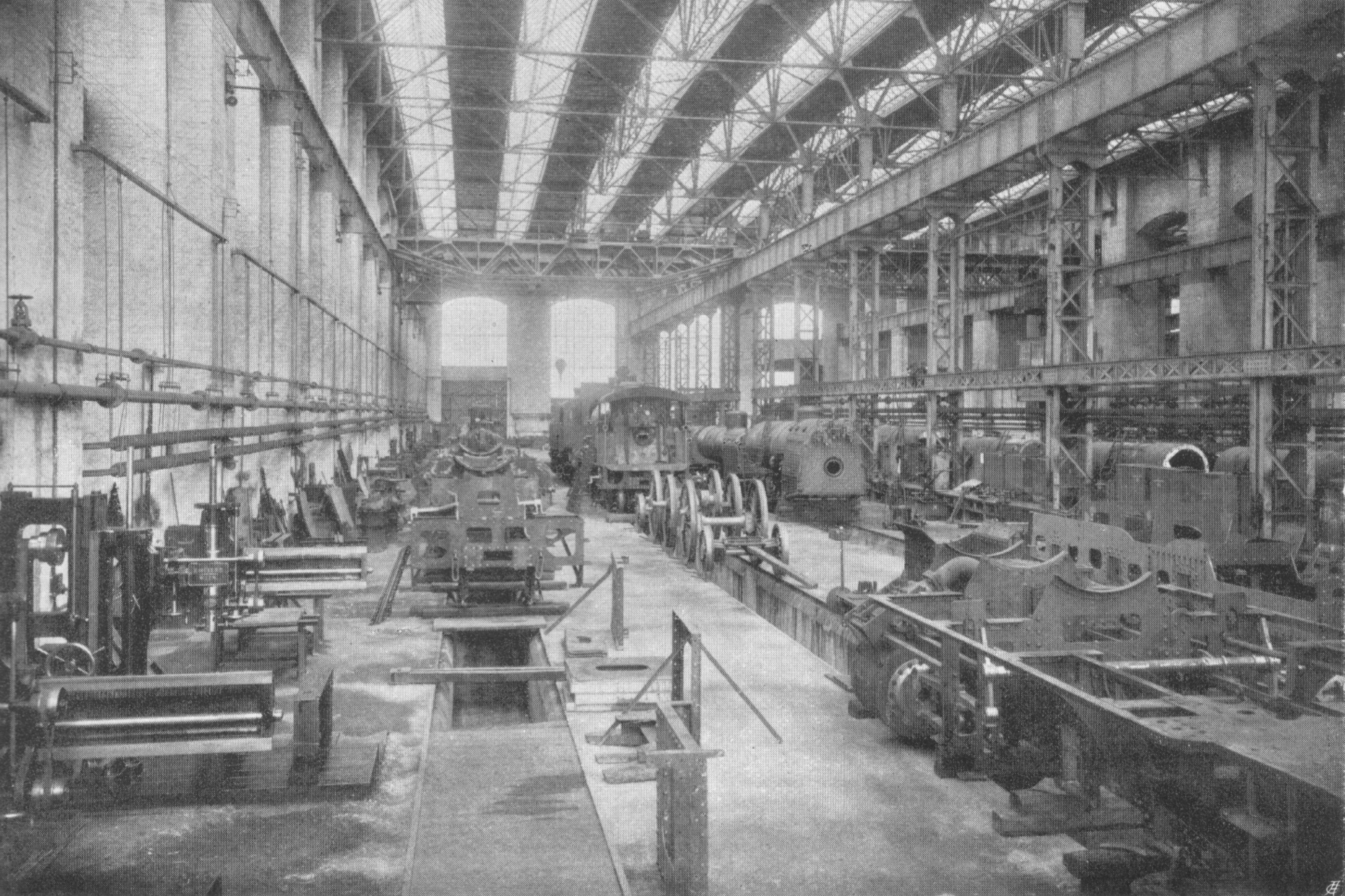 Schwarzes und weißes Foto einer großen Fabrikhalle mit vielen Zügen auf dem Boden, Maschinen, Stützpfosten, Wänden und einer sichtbaren Dachstruktur.