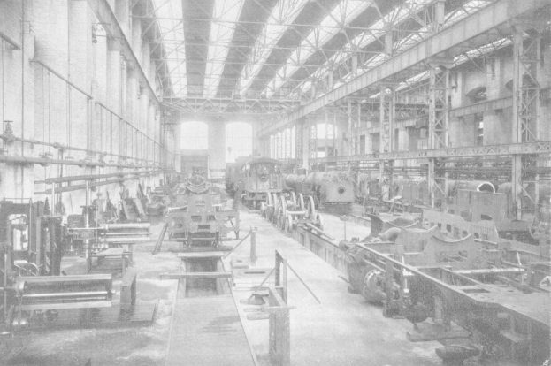 Schwarzes und weißes Foto einer großen Fabrikhalle mit vielen Zügen auf dem Boden, Maschinen, Stützpfosten, Wänden und einer sichtbaren Dachstruktur.