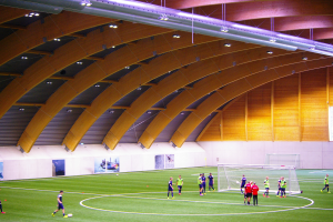 Eine Gruppe von Menschen, die Fußball auf einem grünen Feld in einer großen Halle mit Torpfosten, Wandplakaten und hellen Deckenlampen spielen.