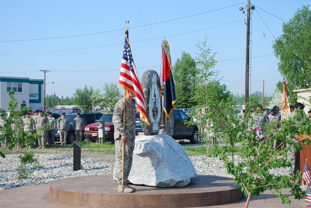 Zwei Militärangehörige stehen mit Fahnen auf beiden Seiten eines Gedenksteins, mit Pflanzen, Autos, anderen Militärangehörigen, Strommasten mit Kabeln und Gebäuden im Hintergrund.