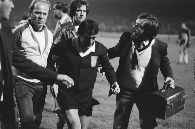 Schwarzes und weißes Bild einer Gruppe von Männern, die auf einem Fußballfeld gehen, einer hält eine Aktentasche, mit offensichtlicher Besorgnis.