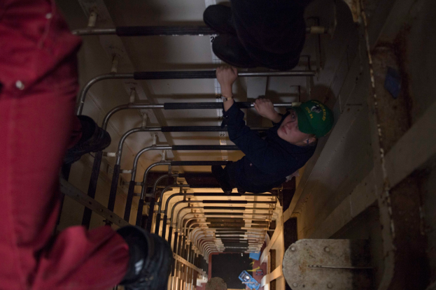 Ein Mann mit grüner Mütze klettert eine Leiter in einem Gebäude hoch, umgeben von Stangen und anderen Gegenständen, trägt Schuhe.