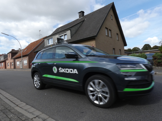 Schwarzer und grüner Skoda Kodiaq elektrischer SUV auf einer Stadtstraße mit Gebäuden, Bäumen und bewölktem Himmel im Hintergrund.