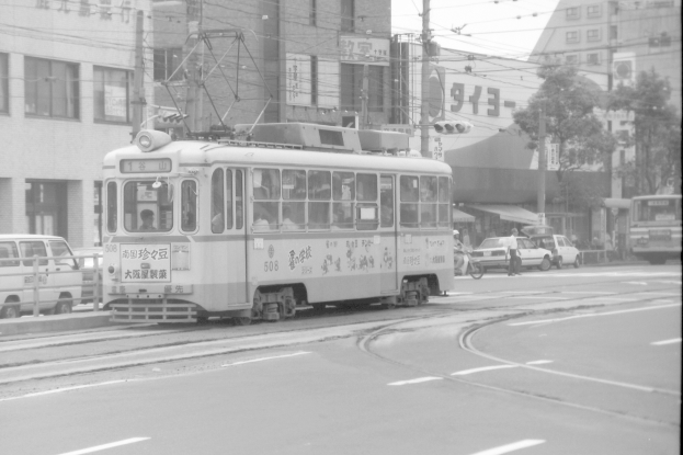 Ein Schwarz-Weiß-Foto einer Stadtstraße mit einer Tram, Fahrzeugen, Radfahrern, Gebäuden mit Fenstern und Strommasten mit Drähten.