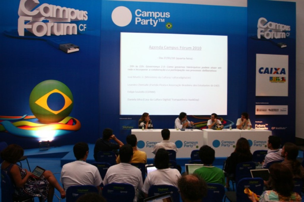 Ein Konferenzsaal mit Menschen an Tischen mit Laptops und Gläsern Wasser, ein Projektionsschirm mit Inhalt und eine blaue Tafel mit Text auf der rechten Seite.