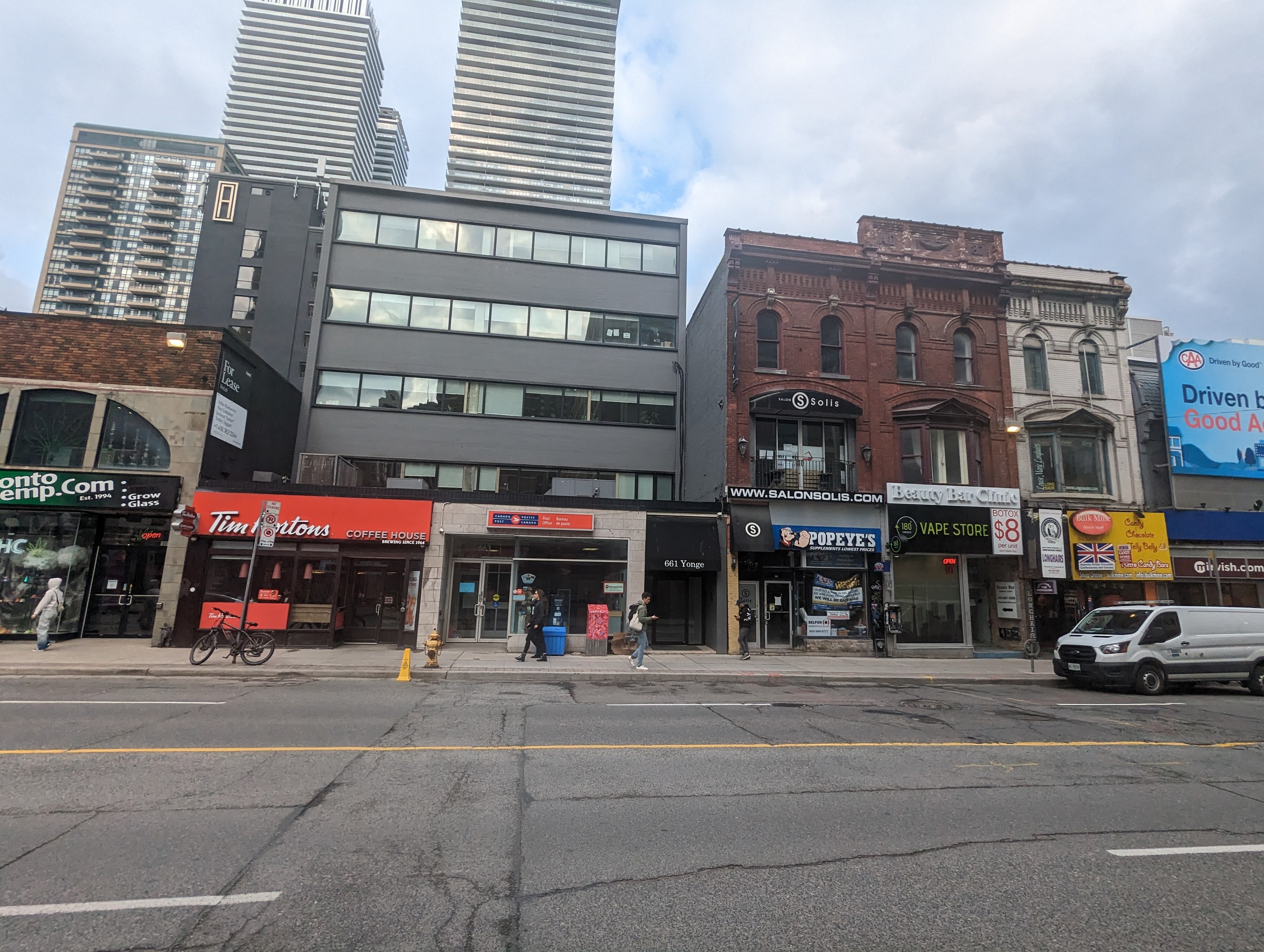 Stadtstraße mit hohen Gebäuden, Fahrzeugen, Fußgängern, einem Fahrrad, einem Feuerhydranten und Werbetafeln für Vermietungen unter einem bewölkten Himmel.