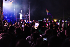 Menschenmenge auf einem Konzert mit hochgehaltenen Handys, beleuchtet von Scheinwerfern, umgeben von Pfählen und einer Fahne, während Musiker auf der Bühne spielen.