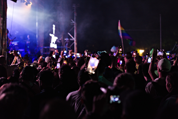 Menschenmenge auf einem Konzert mit hochgehaltenen Handys, beleuchtet von Scheinwerfern, umgeben von Pfählen und einer Fahne, während Musiker auf der Bühne spielen.