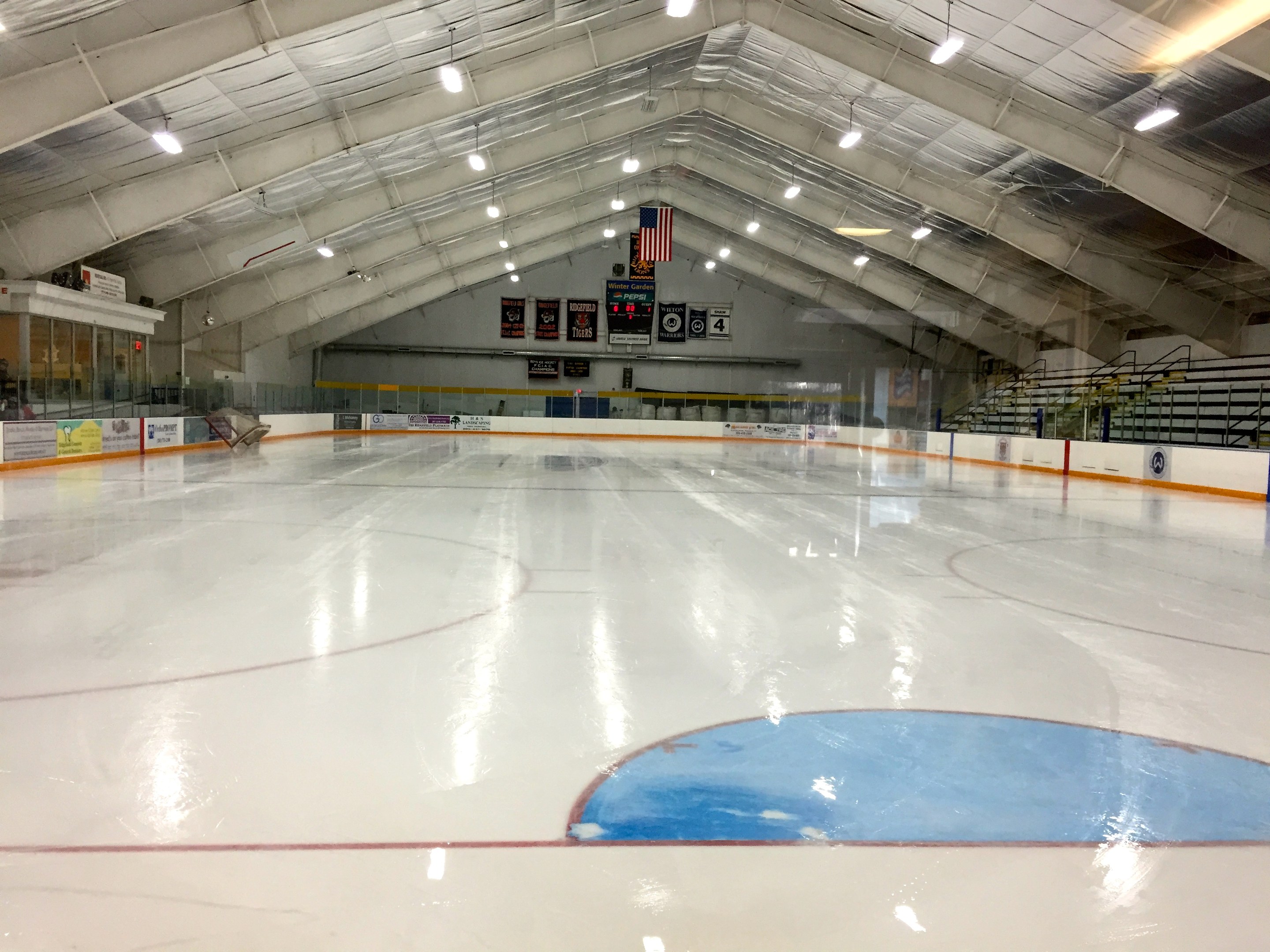 Großes Hallen-Eisstadion mit Deckenbeleuchtung, Werbetafeln, einer Rückwand, Treppen mit Geländern und einigen Menschen, was auf eine Sportanlage hinweist, die likely für Hockey genutzt wird.