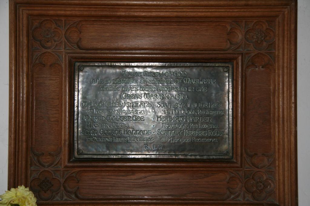 Eine Gedenktafel mit Text ist an einer braunen Wand oder einer hölzernen Tür angebracht, mit einem gelben Blumentopf in der linken unteren Ecke.