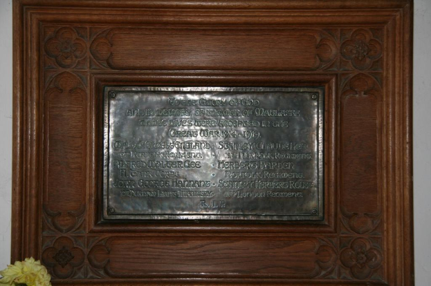 Eine Gedenktafel mit Text ist an einer braunen Wand oder einer hölzernen Tür angebracht, mit einem gelben Blumentopf in der linken unteren Ecke.