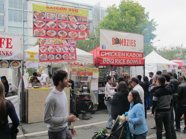 Menschen, die auf einer Straße vor der Stadt stehen, mit einem Mann in einem grauen und weißen T-Shirt, der eine Wasserflasche hält, und einer Frau in einer blauen Jacke, die einen Kinderwagen schiebt. Im Hintergrund sind Zelte in Weiß und Rot, farbenfrohe Banner und Gebäude, Bäume und eine Straßenlaterne zu sehen.