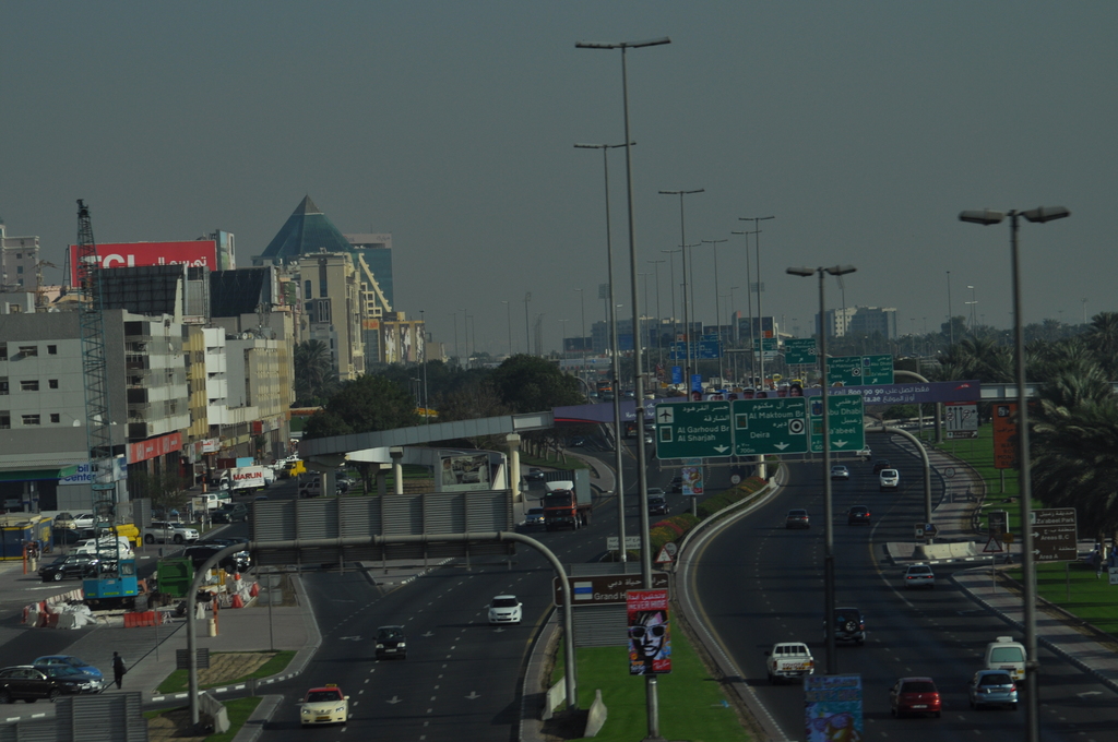 Eine Luftaufnahme einer Stadt mit Gebäuden, Türmen, Kraftfahrzeugen, Werbetafeln, Schildern, Straßen, Bäumen, Straßenlaternen, Menschen, Absperrgittern und Informationsanzeigen unter einem klaren Himmel.