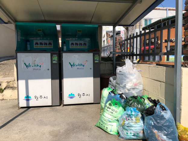 Zwei Getränkeautomaten nebeneinander auf einer Straße mit Polyethylenabdeckungen davor, einem Geländer und einer Wand rechts sowie Gebäuden, Pfählen, Drähten und Himmel im Hintergrund.