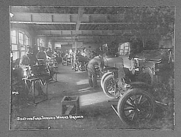 Schwarzes und weißes Foto von Männern, die an Autos in einer Ford-Werkstatt arbeiten, mit Fahrzeugen, Werkzeugen auf Tischen und Fenstern links.