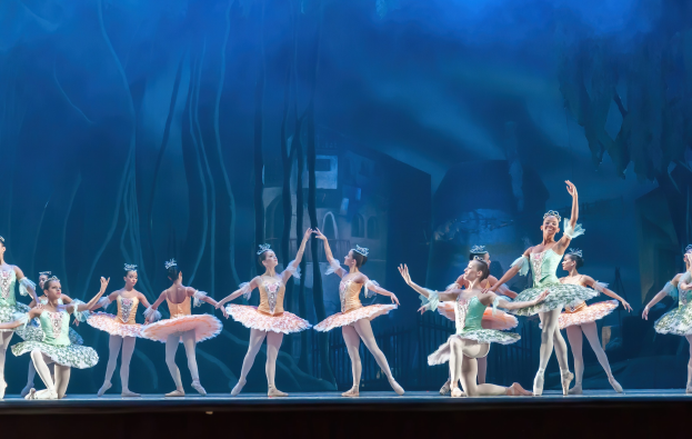 Eine Gruppe von Balletttänzern in farbenfrohen Kostümen, die *Der Nussknacker* auf der Bühne des Royal Opera House tanzen, synchronisiert und anmutig gegen einen blauen Hintergrund.