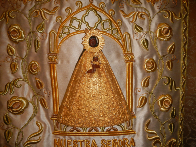Goldbestickte Chasuble mit einer friedlichen Frau in einem hochgeschlossenen, langärmeligen Kleid mit einem Dutt, umgeben von detaillierten Designs und prächtigem Text in einem goldenen Rahmen.